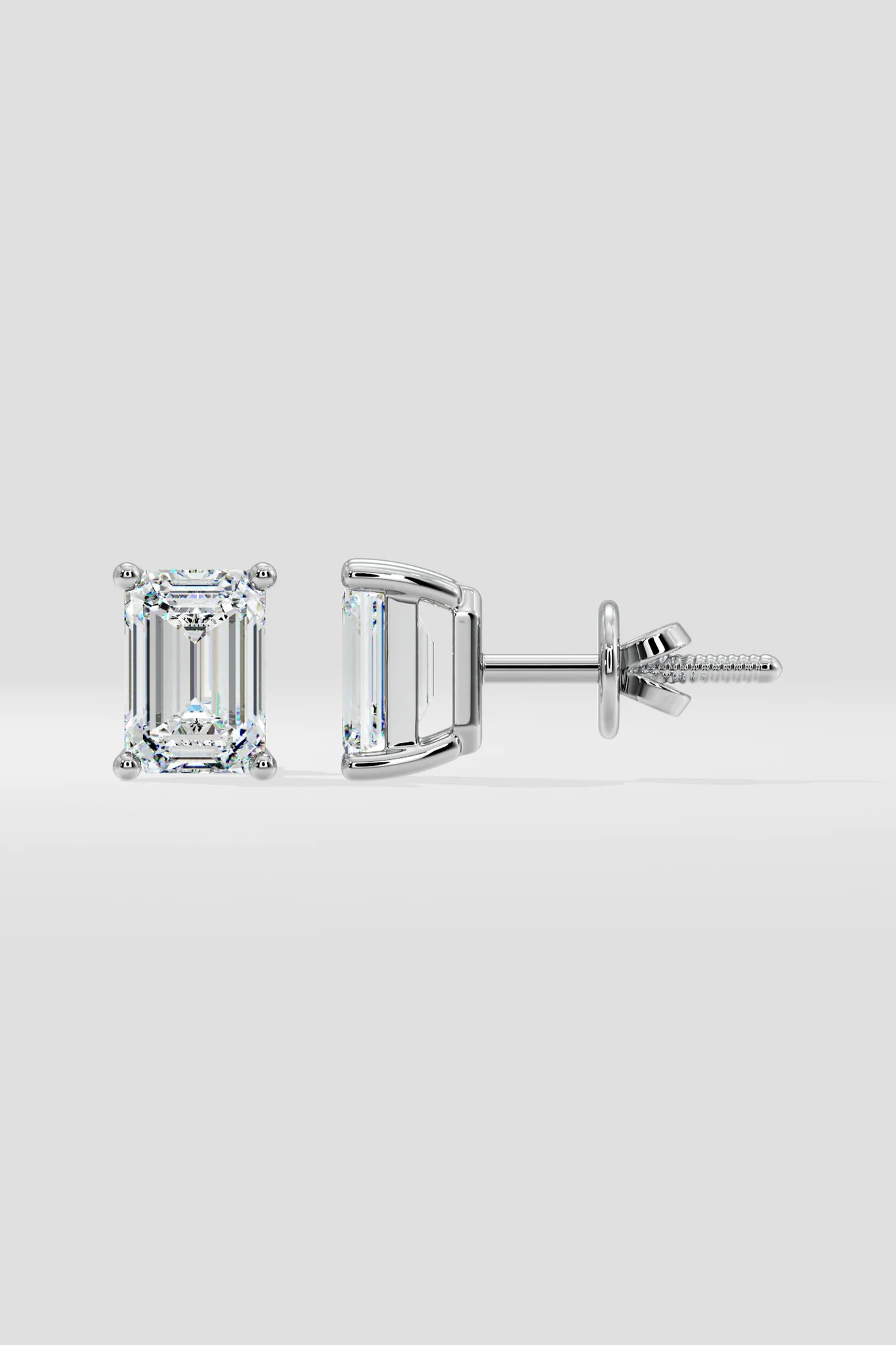 The EMERALD SOLITAIRE EARRINGS are a pair of emerald-cut lab-grown diamond studs with VS2 clarity, set in white metal prongs and displayed from the front and side on a plain light gray background.
