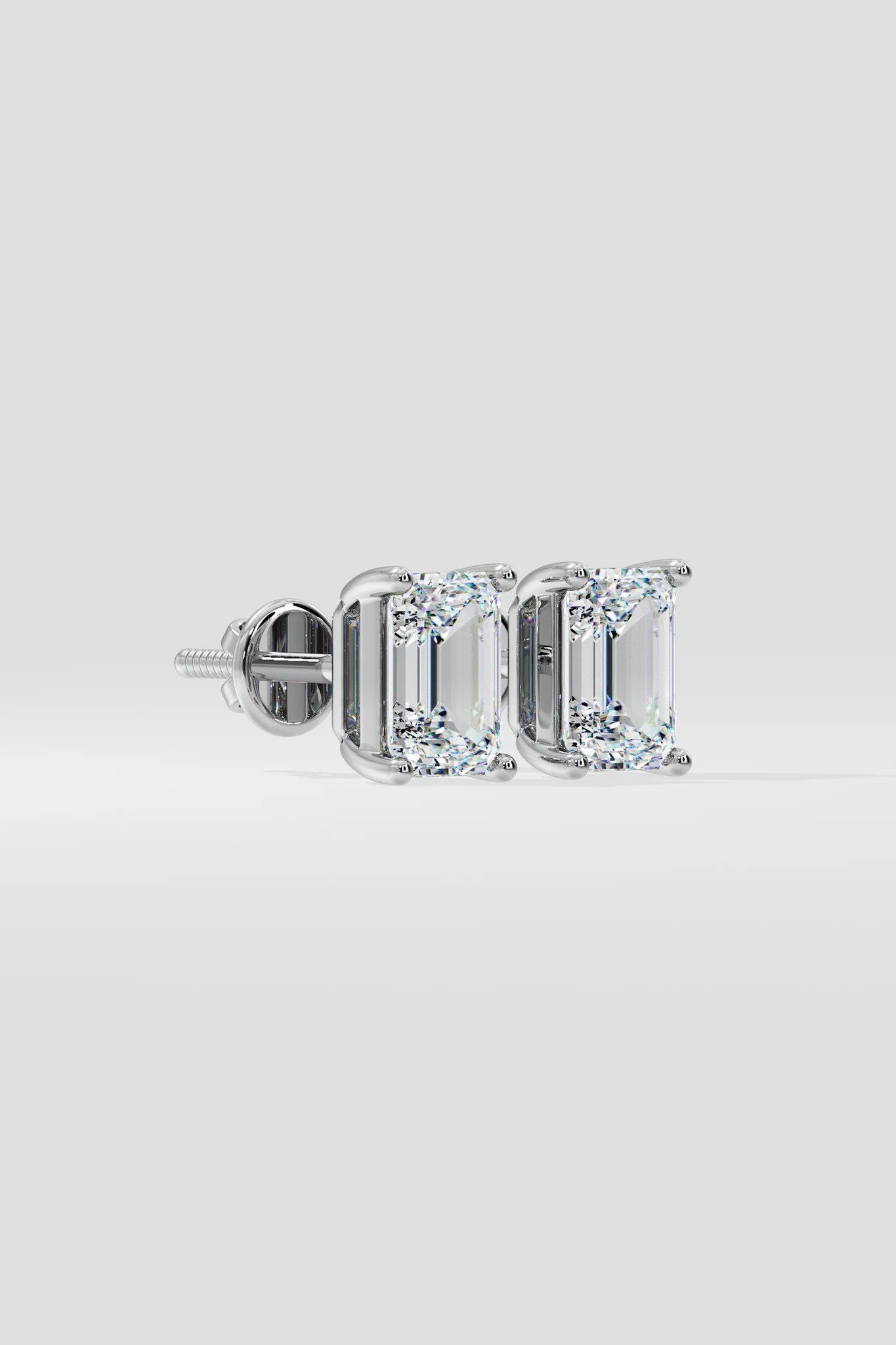 The EMERALD SOLITAIRE EARRINGS are lab-grown, emerald-cut diamond studs with VS2 clarity set in silver prongs, shown on a light gray background.