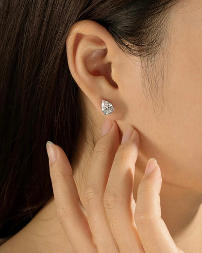 A close-up shows a person with straight, dark hair wearing PEAR SOLITAIRE EARRINGS—large, pear-shaped lab-grown diamonds of VS2 clarity. One hand gently touches the earlobe, showcasing the sparkling jewelry.