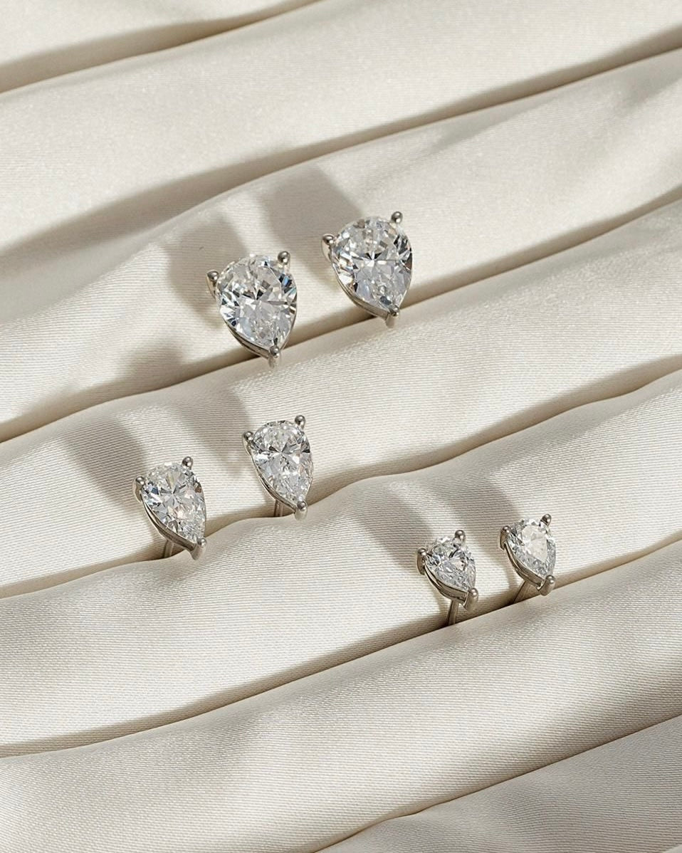Five PEAR SOLITAIRE EARRINGS, each featuring pear-shaped lab-grown diamonds with VS2 clarity in various sizes, are arranged in rows on gently folded, textured cream fabric.