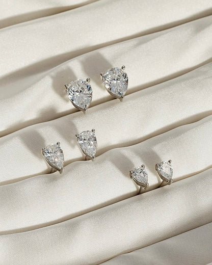 Five PEAR SOLITAIRE EARRINGS, each featuring pear-shaped lab-grown diamonds with VS2 clarity in various sizes, are arranged in rows on gently folded, textured cream fabric.