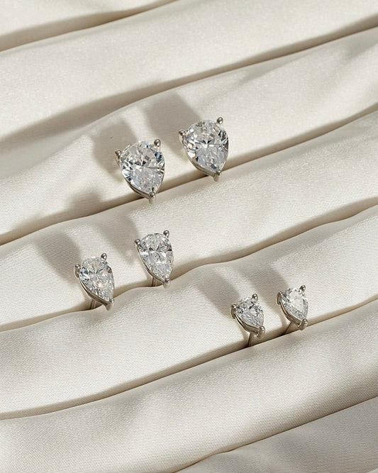 Five PEAR SOLITAIRE EARRINGS, each featuring pear-shaped lab-grown diamonds with VS2 clarity in various sizes, are arranged in rows on gently folded, textured cream fabric.