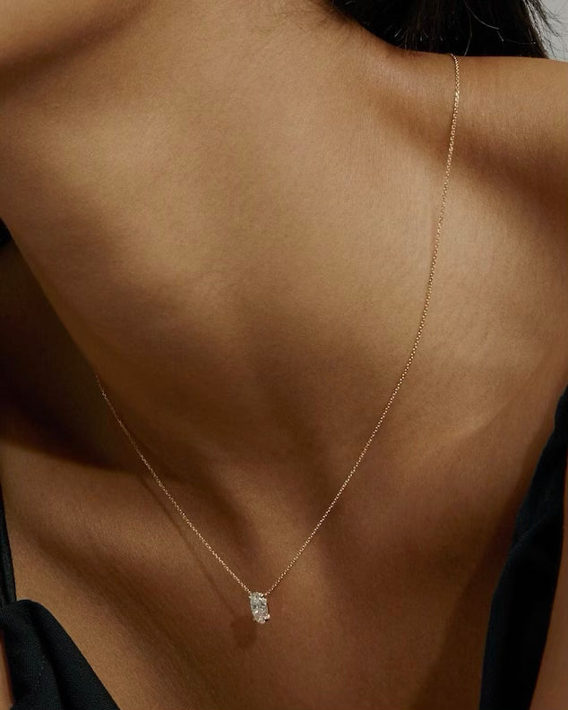 A woman in a black top wears the OVAL SOLITAIRE NECKLACE—a thin gold oval chain with a sparkling lab grown diamond pendant of VS2 clarity. Soft lighting highlights her neck and upper chest.