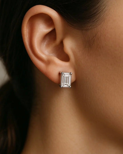 Close-up of a person wearing EMERALD SOLITAIRE EARRINGS featuring a rectangular, lab-grown diamond in a simple metal prong setting. The VS2 clarity stone sparkles against the neutral background, with hair pulled back.
