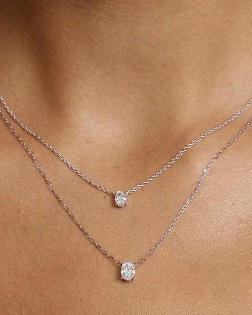 A close-up of a person wearing two delicate silver OVAL SOLITAIRE NECKLACES, each with a small oval pendant featuring a clear lab-grown diamond, layered above the collarbone.